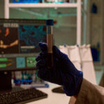 Gloved hand holding a blood sample in a modern medical lab, with digital monitors in the background.